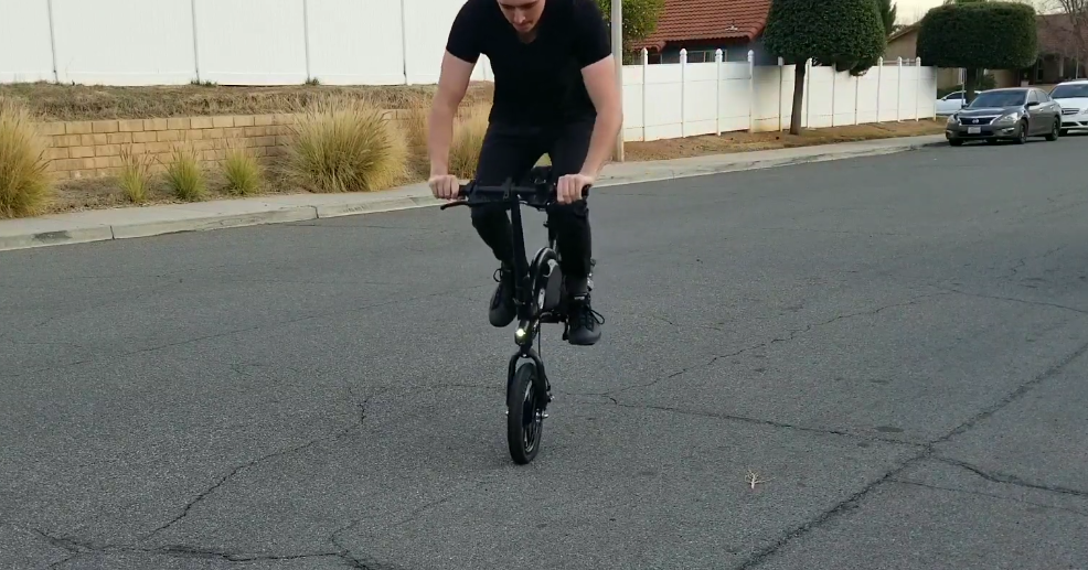Man performing a wheelie on a compact folding electric bike on a suburban street, showcasing agility and fun riding style.