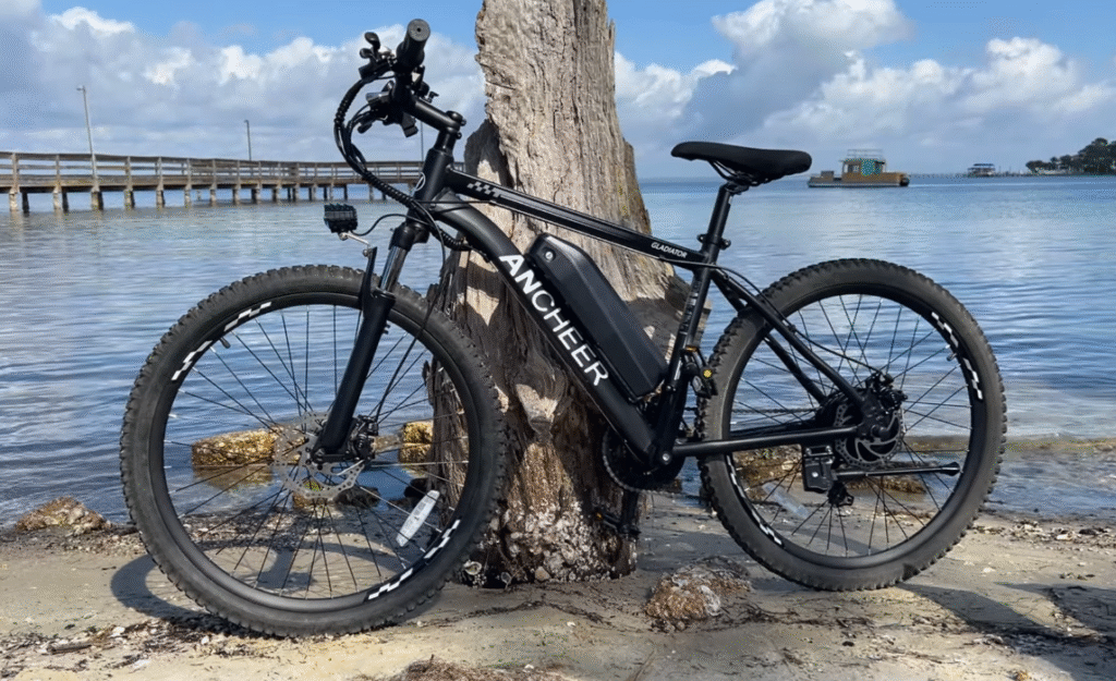 Ancheer Electric Bike Review 2 Side profile of an Ancheer Gladiator electric mountain bike near a seawall, highlighting front suspension fork and drivetrain.