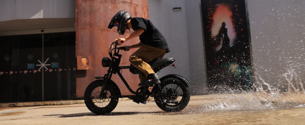 Rider on a black electric bike splashing through water in an urban setting, wearing a helmet.