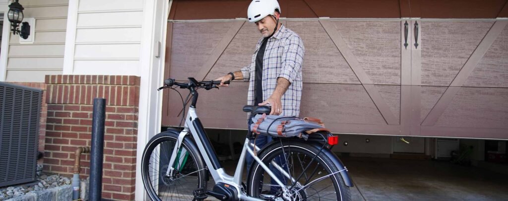 Man preparing to ride a Haven electric bike outside his garage, wearing a helmet and carrying a commuter bag on the rear rack.