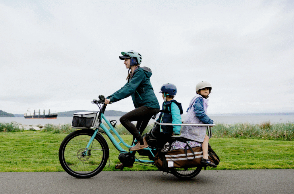 Woman riding a long-tail cargo e-bike with two kids near the seaside.