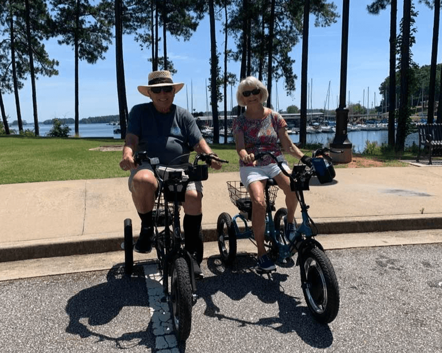 Elderly couple riding electric tricycles by a lakeside park.