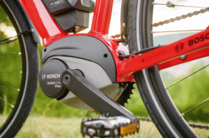 Red e-bike frame with a Bosch Active Line Plus mid-drive motor installed, connected to a chain drivetrain, pedals, and rear wheel, set against a grassy outdoor background.
