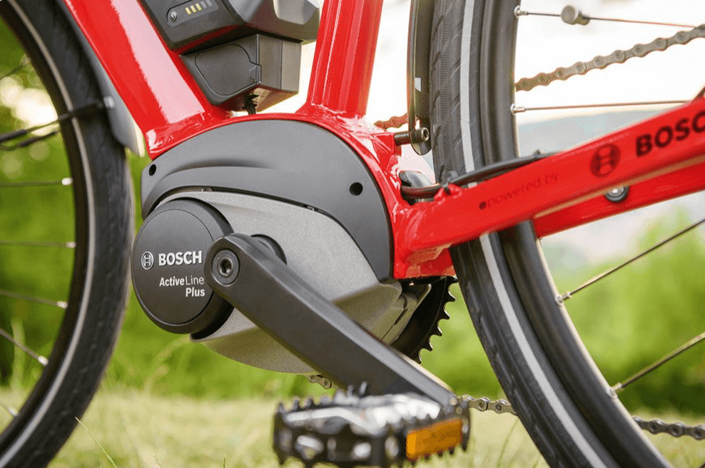Pedal "Clunk" at Start on Belt-Drive E-Bikes: All Likely Causes & Fixes 4 Red e-bike frame with a Bosch Active Line Plus mid-drive motor installed, connected to a chain drivetrain, pedals, and rear wheel, set against a grassy outdoor background.
